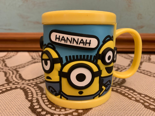 Vintage yellow acrylic child’s mug with Despicable Me Minion Mayhem design, covered in playful Minions and personalized with “Hannah” on one side; Universal Studios logo printed prominently on one side.