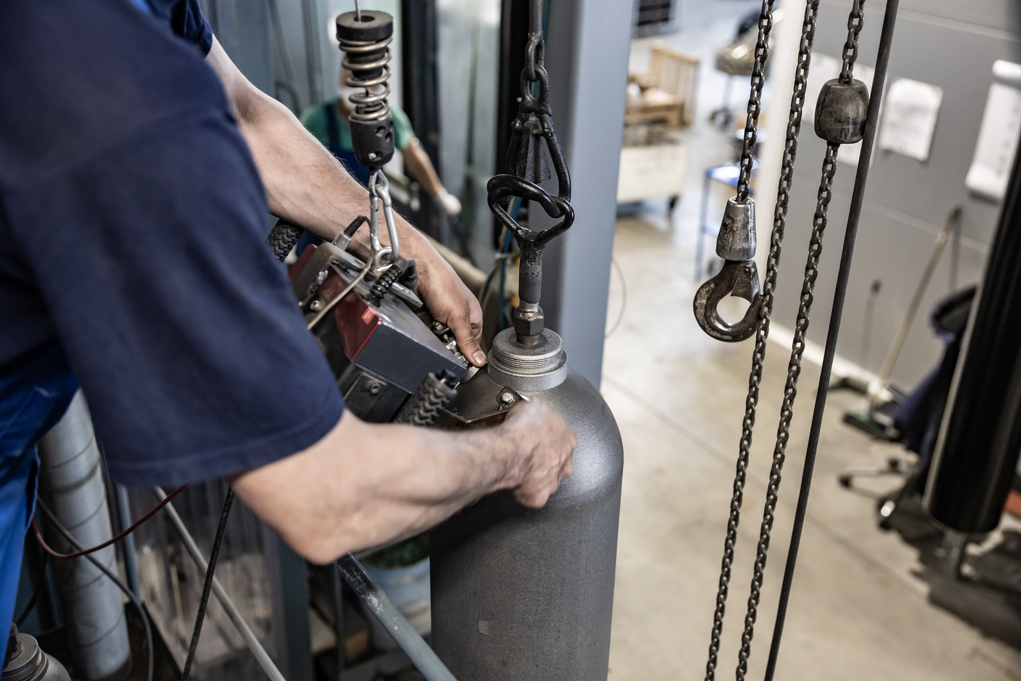 Technician inspecting and testing an industrial gas cylinder using lifting equipment in a workshop.
