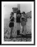 Historic Framed Print, [Mack Sennett comedy films. glamour pose by young woman in bathing suit on beach blowing into meter measuring her breath and man standing alongside],  17-7/8" x 21-7/8"