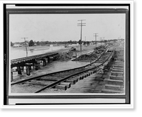 Railroad tracks, one of which is suspended in air, by ground weight on both sides, its support washed away by floodwaters, partially submerged buildings in background.Ewing, Inc. Timeless Wall Ar