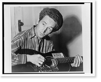 Historic Framed Print, [Woody Guthrie, half-length portrait, seated, facing front, playing a guitar that has a sticker attached reading: This Machine Kills Fascists].World Telegram photo by Al Aumuller.,  17-7/8" x 21-7/8"