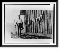 Man, back to water, holding rod and reel, several fish strung up on exterior of wooden building, Vintage Print