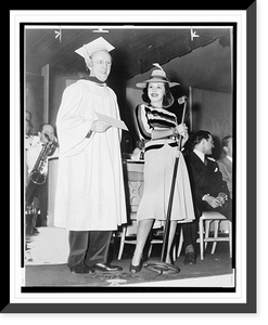 Historic Framed Print, [Kay Kyser, wearing cap and gown, holding a diploma(?), standing next to a woman holding a microphone, with his orchestra in the background],  17-7/8" x 21-7/8"