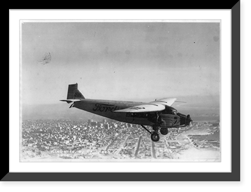Historic Framed Print, [Closeup view of Ford Trimotor plane in flight over unknown city],  17-7/8" x 21-7/8"