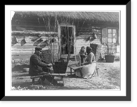 Man and woman making baskets outdoors as small child and others in doorway watch, Poland, Vintage Print