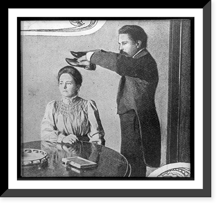 Seance with man holding pair of shoes over head of woman, who is seated at table, Vintage Print