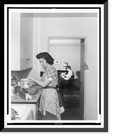 Woman beating eggs in kitchen, and man seated at desk in background, Greenbelt, Maryland, Historic Print