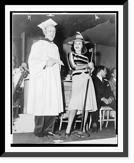 Historic Framed Print, [Kay Kyser, wearing cap and gown, holding a diploma(?), standing next to a woman holding a microphone, with his orchestra in the background],  17-7/8" x 21-7/8"
