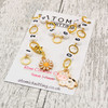 Atomic Knitting row counter chain with gold rings, white acrylic number beads (1-9), and adorable charms featuring a daisy and a cute cow. Displayed on a branded card, perfect for keeping track of knitting or crochet rows in style.