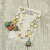 Atomic Knitting row counter chain featuring gold rings, white acrylic number beads (1-9), and square czech beads with colourful patterns. Accented with sparkling beads, the chain is displayed on a branded card on a rustic wood background.