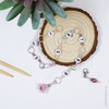 Pink and purple beaded chain row counter set for knitting, featuring white numbered beads from 1 to 9, silver rings, and a pink flower charm, displayed on bamboo knitting needles on a wood slice on a white background.