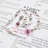 Pink and purple beaded chain row counter set for knitting, featuring white numbered beads from 1 to 9, silver rings, and a pink flower charm, displayed on bamboo knitting needles on a knitting pattern background.