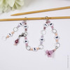 Pink and purple beaded chain row counter set for knitting, featuring white numbered beads from 1 to 9, silver rings, and a pink flower charm, displayed on bamboo knitting needles on a white background.