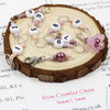 Pink and purple beaded chain row counter set for knitting, featuring white numbered beads from 1 to 9, silver rings, and a pink flower charm, displayed on bamboo knitting needles on a wood slice on a knitting pattern background.
