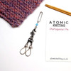 Seahorse Portuguese knitting pin by Atomic Knitting shown beside its white product card displaying atomicknitting.co.uk. The full length of the pin is visible, showing the silver safety pin top, pale blue bead, seahorse charm, lobster clasp and two S-hooks. A pink and purple knitted swatch and wooden double-pointed needles are used as props.