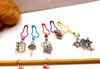 Alice in Wonderland knitting accessory set displayed on white surface with golden skein, showing beaded row counter on needle and five charm stitch markers arranged separately