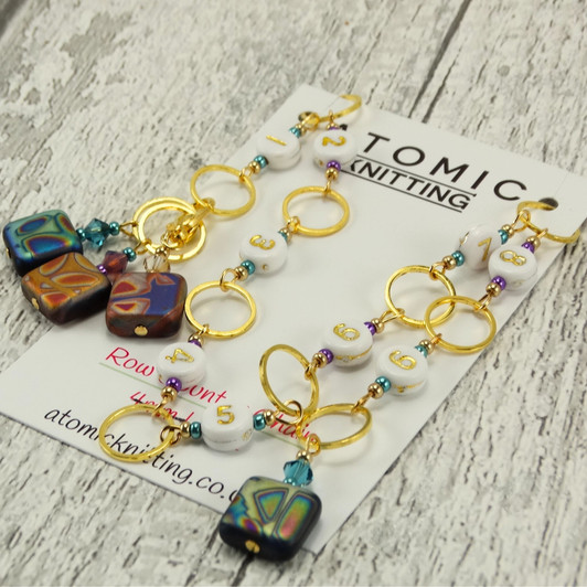 Atomic Knitting row counter chain with gold rings, white acrylic number beads (1-9), and czech beads with colourful patterns. Accented with sparkling beads, the chain is displayed on a branded card on a rustic wood background.