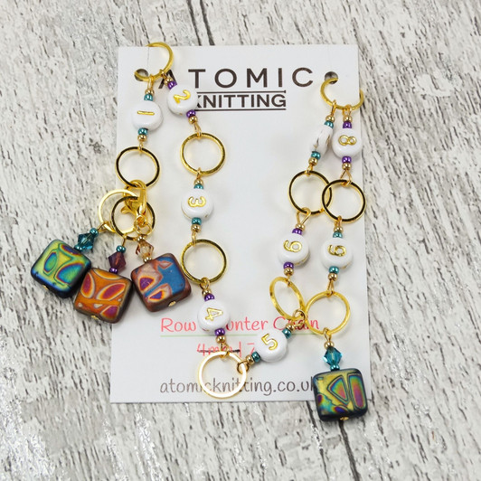Atomic Knitting squares row counter chain featuring gold rings, white acrylic number beads (1-9), and czech beads with colourful patterns. Accented with sparkling beads, the chain is displayed on a branded card on a rustic wood background.