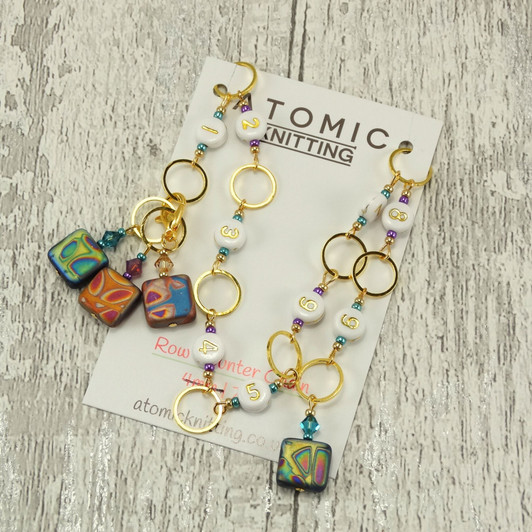 Atomic Knitting row counter chain featuring gold rings, white acrylic number beads (1-9), and square czech beads with colourful patterns. Accented with sparkling beads, the chain is displayed on a branded card on a rustic wood background.