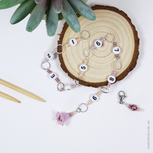 Pink and purple beaded chain row counter set for knitting, featuring white numbered beads from 1 to 9, silver rings, and a pink flower charm, displayed on bamboo knitting needles on a white background.