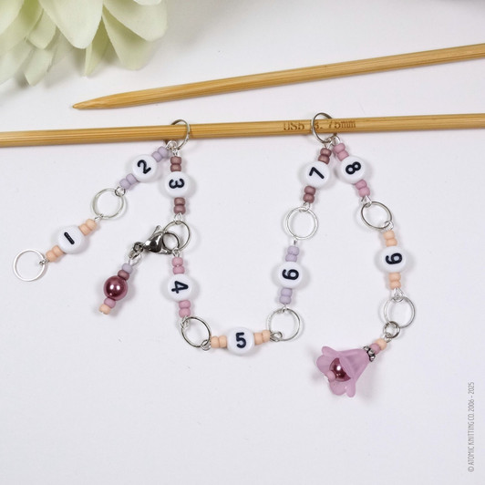 Pink and purple beaded chain row counter set for knitting, featuring white numbered beads from 1 to 9, silver rings, and a pink flower charm, displayed on bamboo knitting needles on a white background.