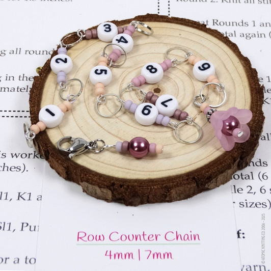 Pink and purple beaded chain row counter set for knitting, featuring white numbered beads from 1 to 9, silver rings, and a pink flower charm, displayed on bamboo knitting needles on a wood slice