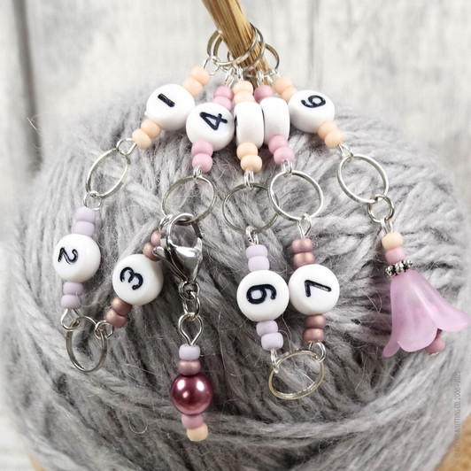 Pink and purple beaded chain row counter set for knitting, featuring white numbered beads from 1 to 9, silver rings, and a pink flower charm, displayed on bamboo knitting needles on a skein of grey yarn.