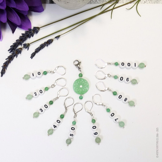 Set of ten green aventurine number stitch markers with silver rings, numbered 10 to 100, displayed with a green pressed glass progress marker, used for knitting and crochet projects.
