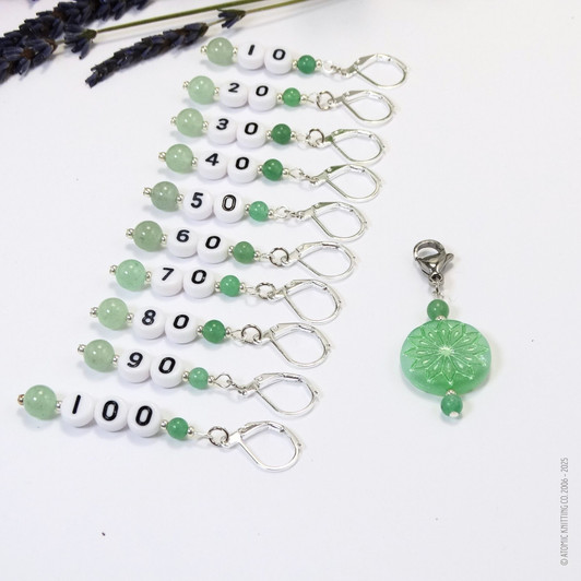Set of ten green aventurine number stitch markers with silver rings, numbered 10 to 100, displayed with a green pressed glass progress marker, used for knitting and crochet projects.