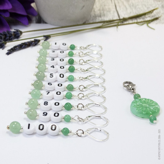 Set of ten green aventurine number stitch markers with silver rings, numbered 10 to 100, displayed with a green pressed glass progress marker, used for knitting and crochet projects.