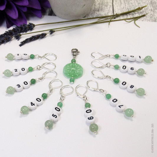 Set of ten green aventurine number stitch markers with silver rings, numbered 10 to 100, displayed with a green pressed glass progress marker, used for knitting and crochet projects.