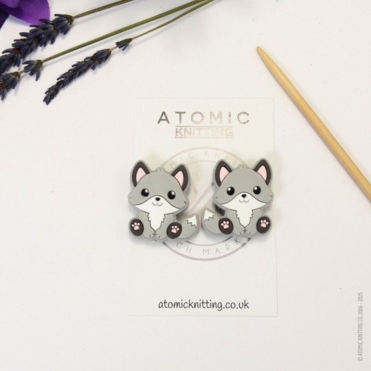 A pair of woodland fox needle stoppers in grey, shaped like cute foxes, placed on the tips of knitting needles to protect points and prevent dropped stitches.