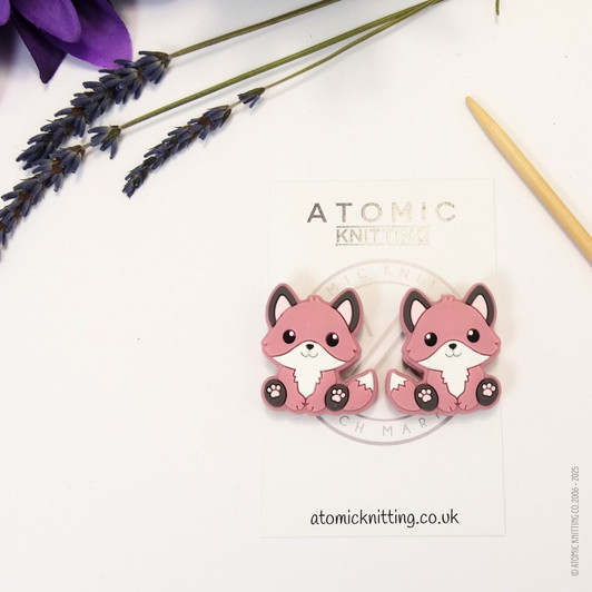 A pair of woodland fox needle stoppers dark pink, shaped like cute foxes, placed on the tips of knitting needles to protect points and prevent dropped stitches.