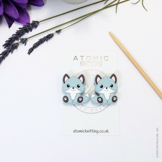 A pair of woodland fox needle stoppers in light blue, shaped like cute foxes, placed on the tips of knitting needles to protect points and prevent dropped stitches.