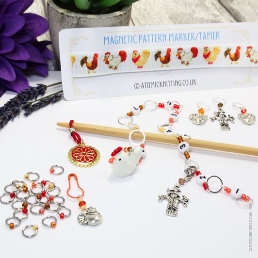 Chicken-themed knitting notions bundle displayed on a white background, including a chicken-print magnetic pattern guide, a beaded row counter chain with number beads and chicken charms, multiple stitch markers with chicken and scarecrow charms, a blunt needle, and orange and red accent beads, all arranged with lavender and a purple flower.