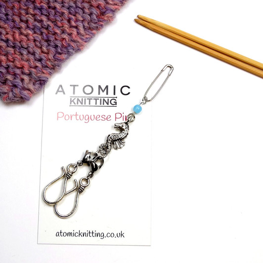 Seahorse Portuguese knitting pin by Atomic Knitting displayed on its white branded product card. The pin features a silver safety pin attachment, a small pale blue bead, a detailed silver seahorse charm, a lobster clasp and two silver S-hooks. A pink and purple knitted swatch and wooden double-pointed needles are visible as styling props.