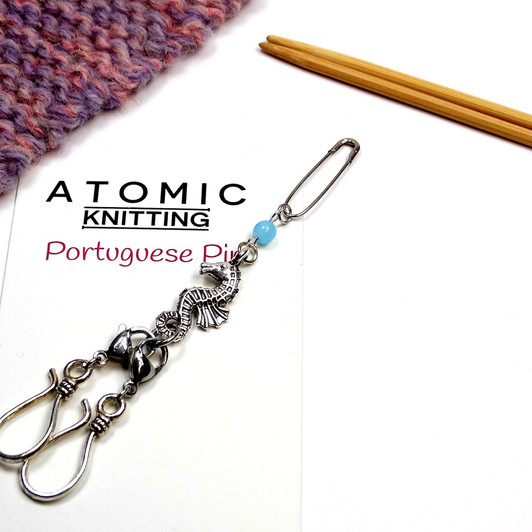 Close-up of a seahorse Portuguese knitting pin by Atomic Knitting laid on its white product card. The silver safety pin, pale blue bead, intricate seahorse charm and paired S-hooks are clearly visible. A pink and purple knitted swatch and wooden knitting needles are arranged in the background.