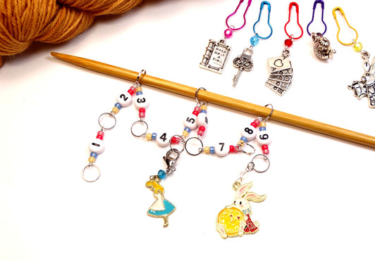 Alice in Wonderland beaded row counter chain threaded on bamboo knitting needle, showing numbers 1–9 with enamel Alice and White Rabbit charms, alongside five silver charm stitch markers