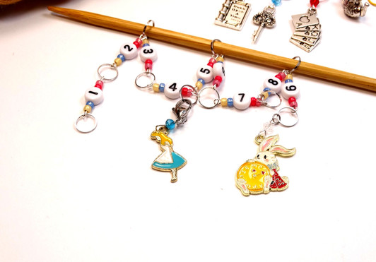 Full flat lay of Alice in Wonderland knitting set: beaded row counter chain and five Alice-themed bulb pin stitch markers arranged on white surface with golden yarn skein