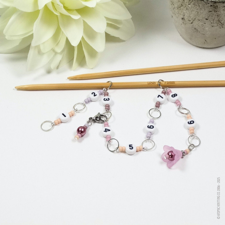 Pink and purple beaded chain row counter set for knitting, featuring white numbered beads from 1 to 9, silver rings, and a pink flower charm, displayed on bamboo knitting needles on a white background.