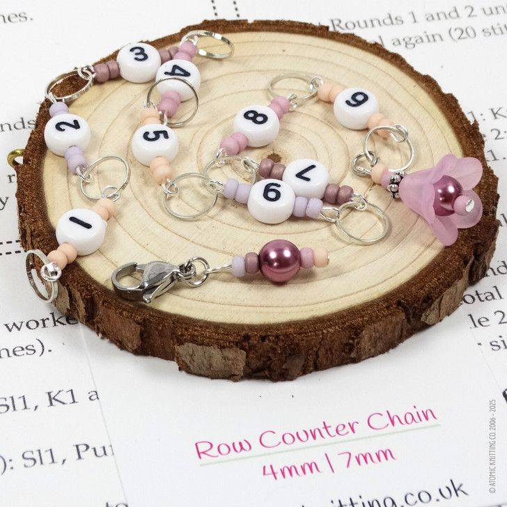 Pink and purple beaded chain row counter set for knitting, featuring white numbered beads from 1 to 9, silver rings, and a pink flower charm, displayed on bamboo knitting needles on a wood slice on a knitting pattern background.