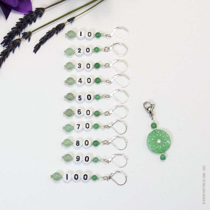 Set of ten green aventurine number stitch markers with silver rings, numbered 10 to 100, displayed with a green pressed glass progress marker, used for knitting and crochet projects.