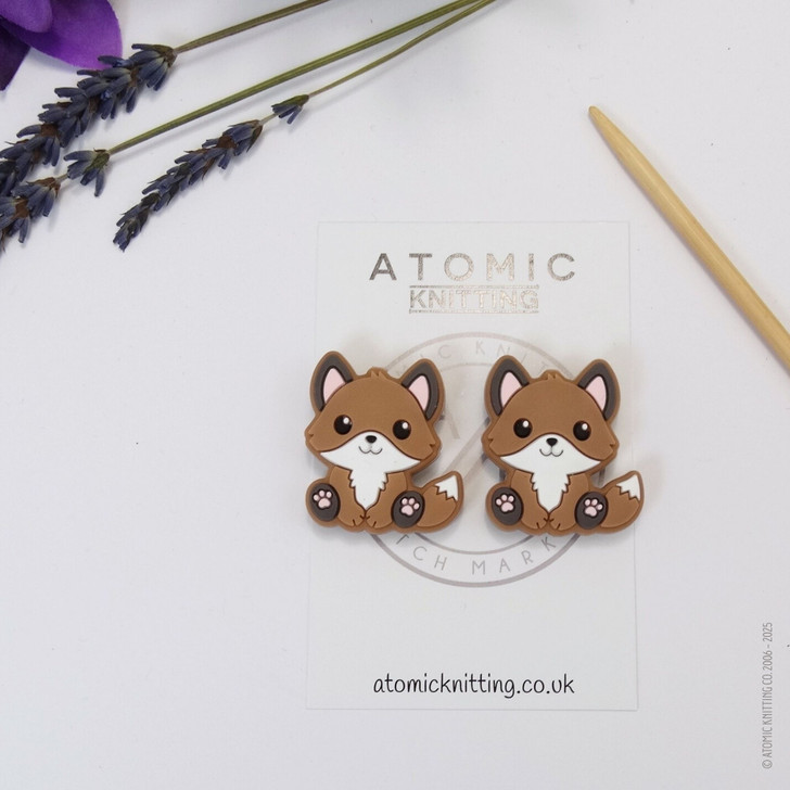 A pair of woodland fox needle stoppers in brown, shaped like cute foxes, placed on the tips of knitting needles to protect points and prevent dropped stitches.