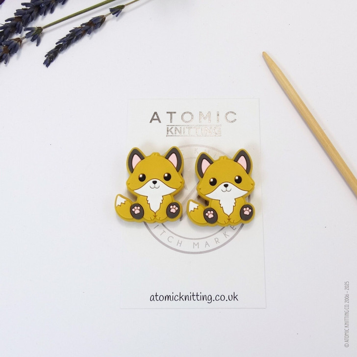 A pair of woodland fox needle stoppers in mustard yellow, shaped like cute foxes, placed on the tips of knitting needles to protect points and prevent dropped stitches.