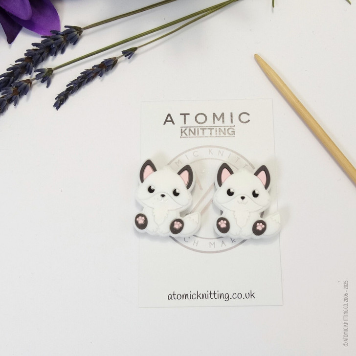 A pair of woodland fox needle stoppers in white, shaped like cute foxes, placed on the tips of knitting needles to protect points and prevent dropped stitches.