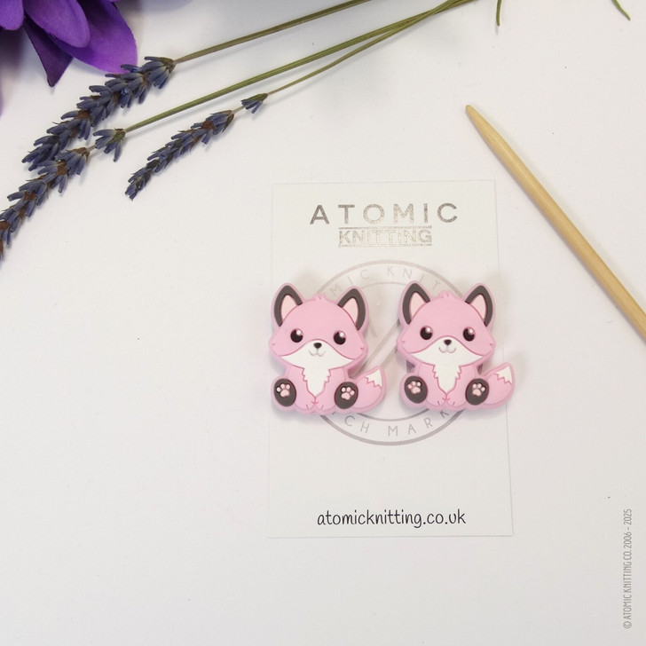 A pair of woodland fox needle stoppers in light pink, shaped like cute foxes, placed on the tips of knitting needles to protect points and prevent dropped stitches.