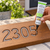 Hand applying Aleene’s Outdoor Adhesive inside a carved 2305 wood sign decorated with rhinestones