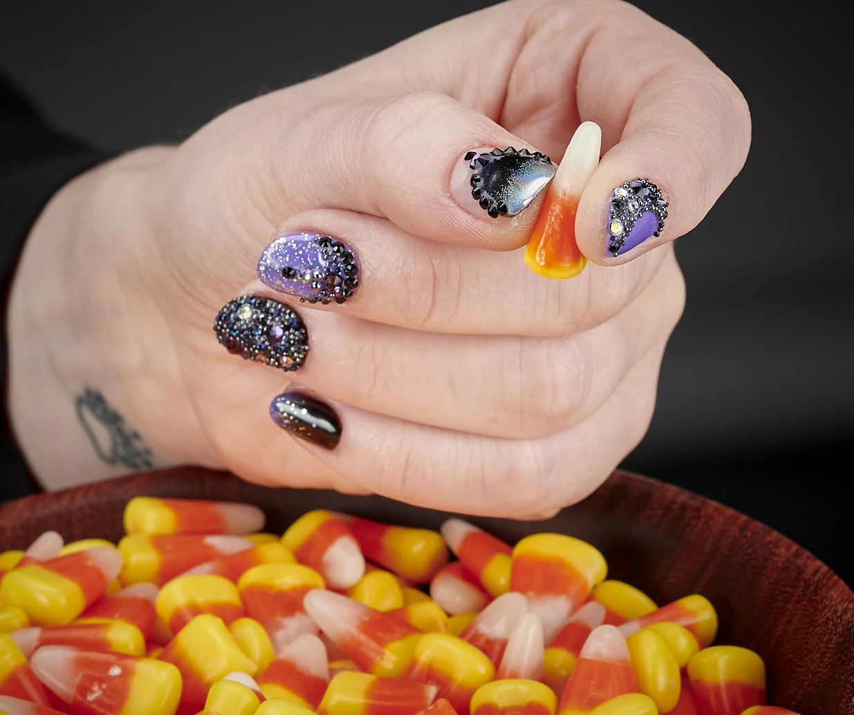Halloween nail art with black and purple crystal detailing and shimmer, shown with a bowl of candy corn