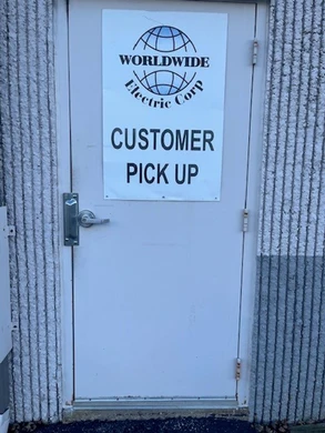 A beige metal door with a white sign reading "WORLDWIDE Electric Corp CUSTOMER PICK UP" in bold text. The door is set in a textured exterior wall, and a silver handle is visible on the left side.