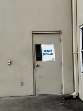 A beige exterior wall with a single tan metal door labeled "Driver Entrance" in large blue letters on a white sign. The door has a small rectangular window, and a downspout is visible to the right of the door. The ground around the door is paved.